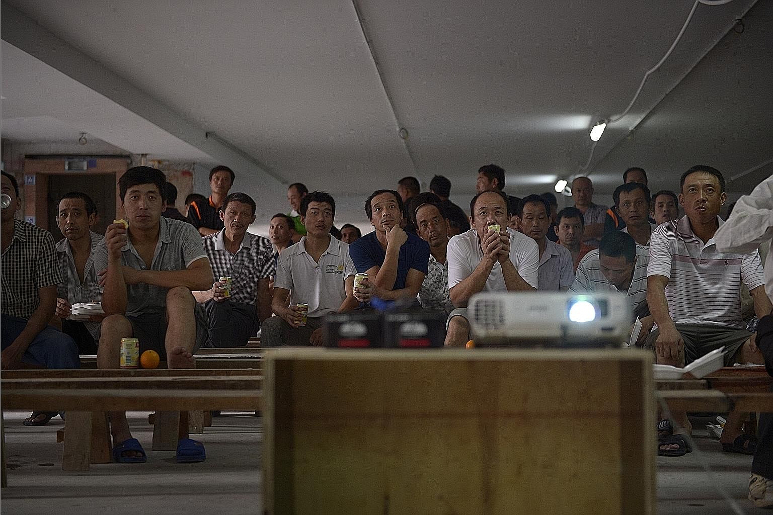 Workers enjoying a movie projected on a makeshift screen inside the multistorey carpark at the Hougang Crimson worksite. Workers finished work early on this day to celebrate the “topping up” of the carpark – a stage of construction when the last structura
