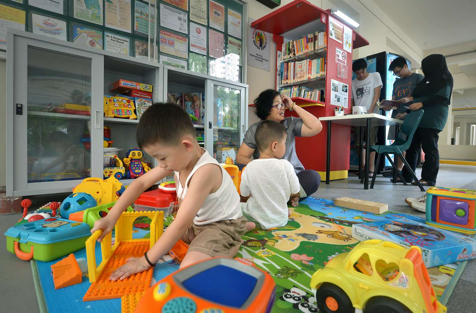 Lee Siang Yu, four, (left) and his brother, Siang Hao, two, with their grandmother, Madam Ho Koon Foong, 61, in the play area of the toy library, while Lim Wee Yang, 16 (standing, in white) and Lai Zi Yang, 16, get help from mobile library assistant Nora 