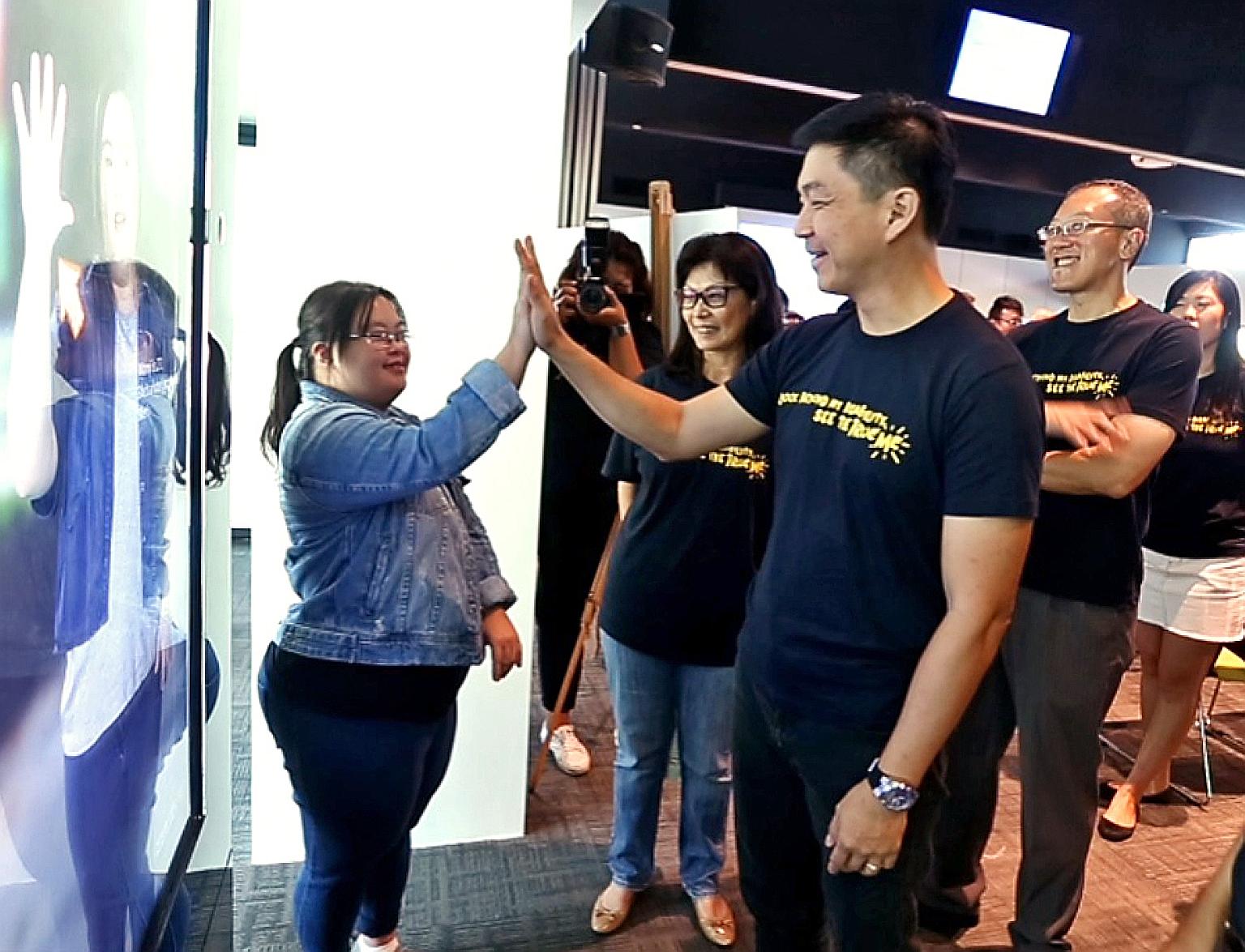 Minister Tan Chuan-Jin exchanging high fives with Ms Chen Wanyi, who has Down syndrome. Behind her is an interactive bus stop ad. If anyone high-fives her image in the ad, she would introduce herself and do a short dance. The bus stop ads are part of