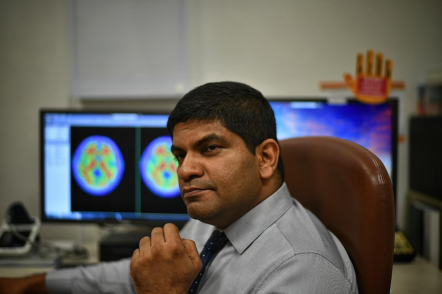 Associate Professor Nagaendran Kandiah in front of brain images showing harmful amyloid plaques that appear in red.