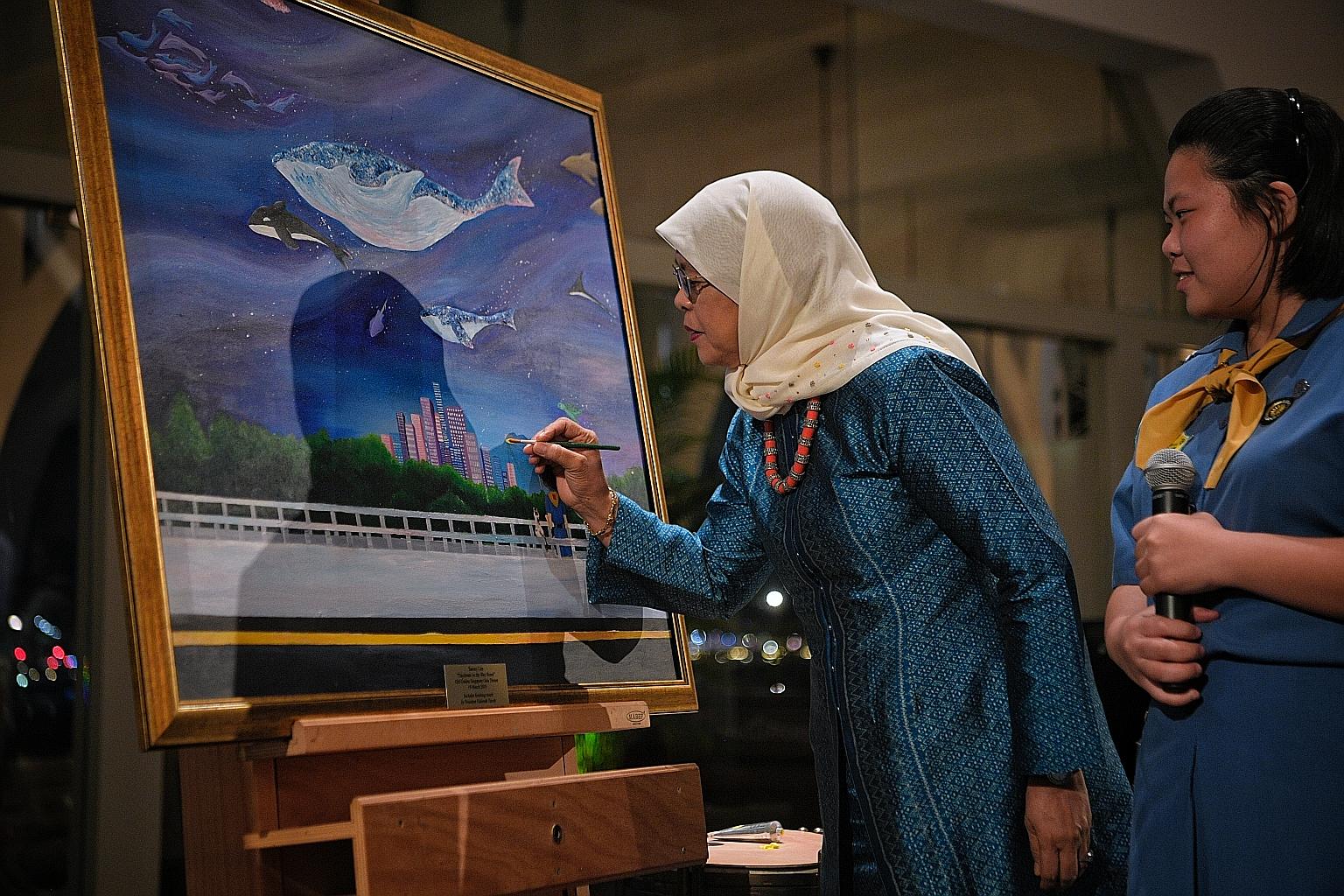 President Halimah Yacob adding a finishing touch to a painting done by Pasir Ris Crest Secondary School student Tammy Lim (right). The painting was one of 12 artworks for sale at yesterday's GGS Gala Dinner.