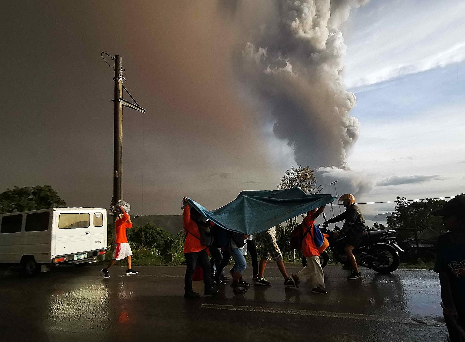 People taking cover under a large plastic sheet as a column of ash spewed from Taal Volcano over Tagaytay city, Philippines, yesterday. Ash fell as far away as Manila, prompting the suspension of flights at the capital's busy international airport. T