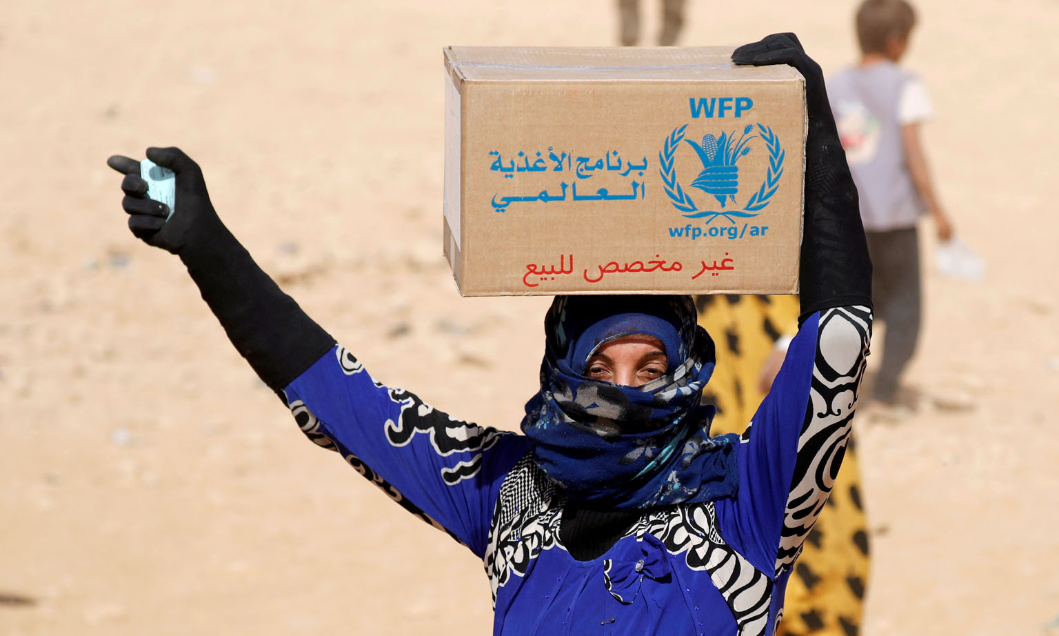 A 2017 photo showing a woman at a refugee camp in Syria holding a food stamp and carrying a box of food aid given by the United Nations' World Food Programme. 