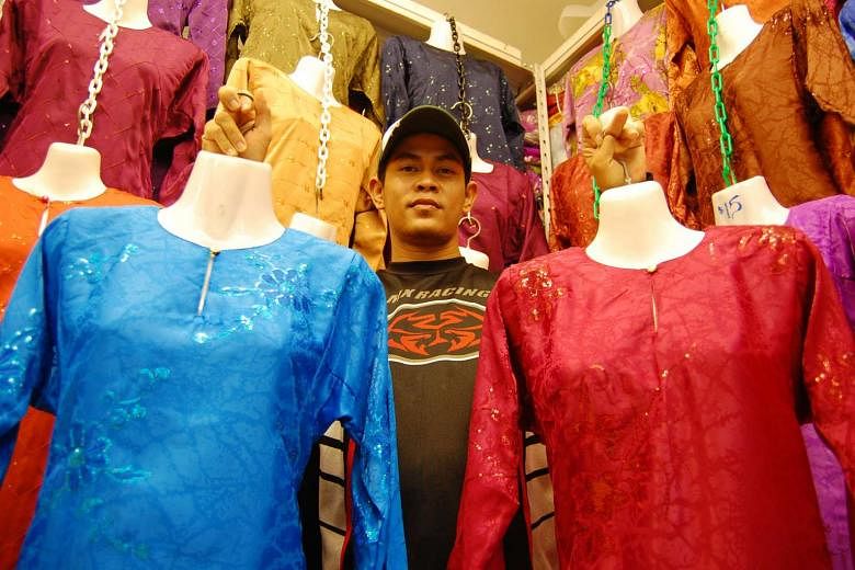 A stallholder in Geylang Serai holding up two traditional Malay costumes which were made in Vietnam.
