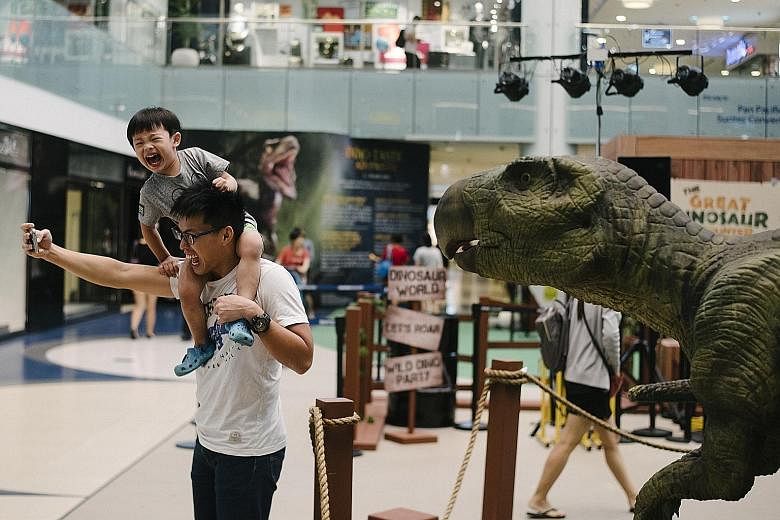 A father and son posing for a selfie with a dinosaur exhibit, which is part of the Dino-Tastic Adventure event held at the Marina Square atrium. The event showcases animatronic dinosaur exhibits as well as a live show, where families get to "feed and