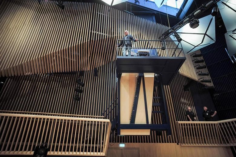 Piano builder David Klavins standing tall next to the 470i Vertical Concert Grand piano in the new concert hall in Ventspils, Latvia. The steel-framed piano hangs as if in mid-air, some three storeys above the audience. To play it, pianists must clim