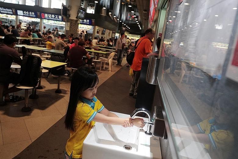 (Far left) The "SG Clean" quality mark at a hawker stall in Our Tampines Hub. (Left) Chua Yu Xuan, seven, washing her hands at the same hawker centre yesterday. Posters have been put up at hawker centres to encourage customers to practise good person
