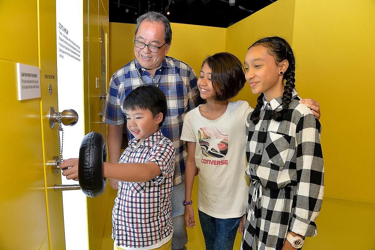 Visitors at Science Centre's Dialogue With Time - Embrace Ageing exhibition unlocking a door while a mechanism keeps their hand shaking, to simulate hand tremors. Mr Victor Lim is in the background.